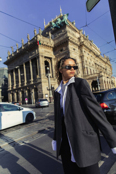 curly african american woman in oversized blazer and sunglasses standing near national theatre in prague