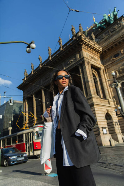 low angle view of young african american woman in stylish blazer and sunglasses posing near national theatre in prague