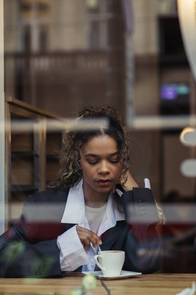 african american woman in trendy blazer stirring coffee in cup while sitting behind window glass in cafe