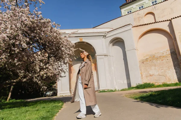 Full length of african american woman in stylish trench coat standing near blooming tree in prague — Photo de stock