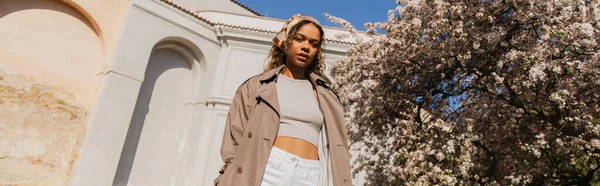 Low angle view of african american woman in stylish outfit standing near blooming tree in prague, banner — Photo de stock