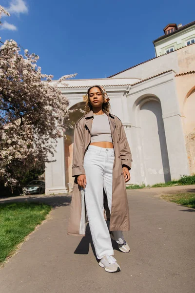 Full length of african american woman in stylish trench coat standing in park near blooming tree — Foto stock