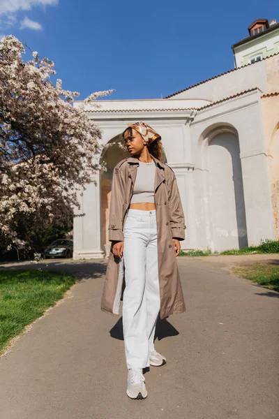 Full length of stylish african american woman in trench coat standing in park near blooming tree — Stock Photo