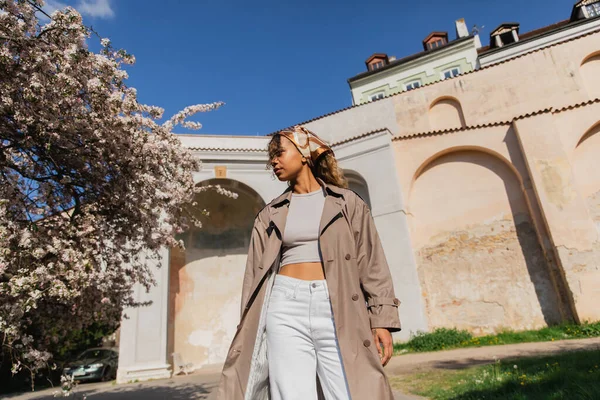 Low angle view of african american woman in trendy outfit standing near blooming tree in prague — Stockfoto