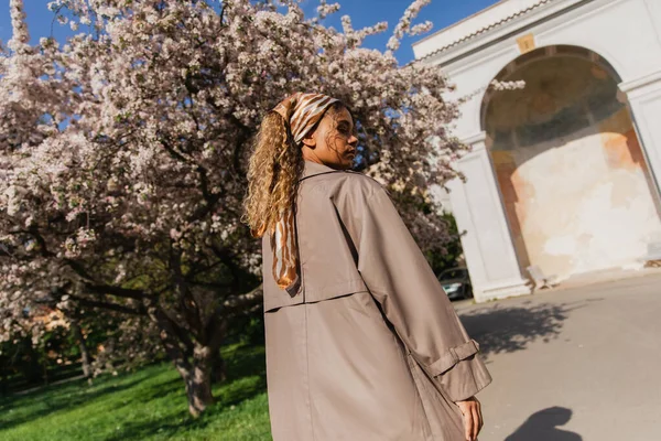 Young african american woman in headscarf and stylish trench coat standing near blooming tree in prague - foto de stock