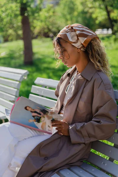 Young african american woman in headscarf and stylish trench coat sitting on bench and reading magazine in prague - foto de stock