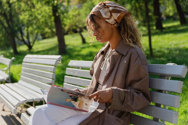 Young african american woman in headscarf and trendy trench coat sitting on bench and reading magazine - foto de stock