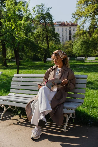 Young african american woman in headscarf and trendy outfit sitting on bench and reading magazine — Fotografia de Stock