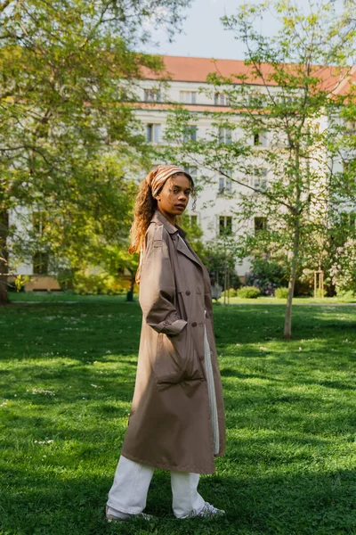 Young african american woman in headscarf and stylish trench coat standing with hand in pocket on green grass in park — Fotografia de Stock