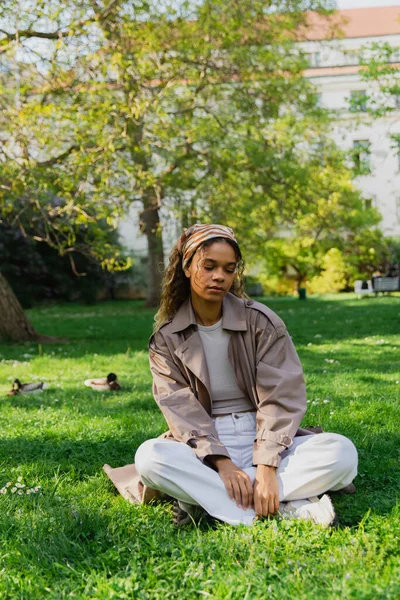 Young african american woman in headscarf and stylish trench coat sitting on green grass in park - foto de stock