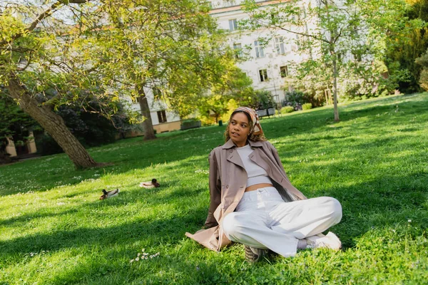 Young african american woman in headscarf and stylish trench coat resting on green grass in park — Stock Photo