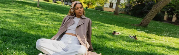 Young african american woman in headscarf and stylish trench coat resting on green grass in park, banner — Stock Photo