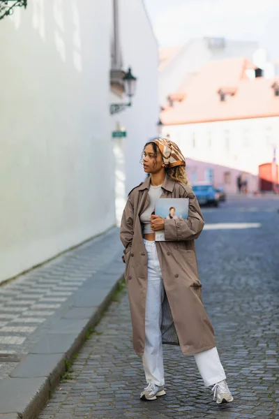 Pretty african american woman in headscarf and trench coat walking with magazine on street in prague - foto de stock