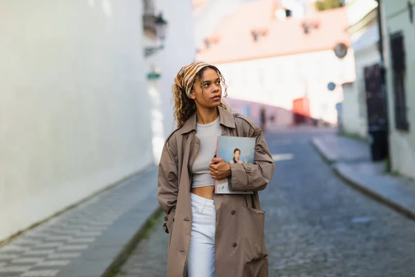 Stylish african american woman in headscarf and trench coat walking with magazine on street in prague - foto de stock