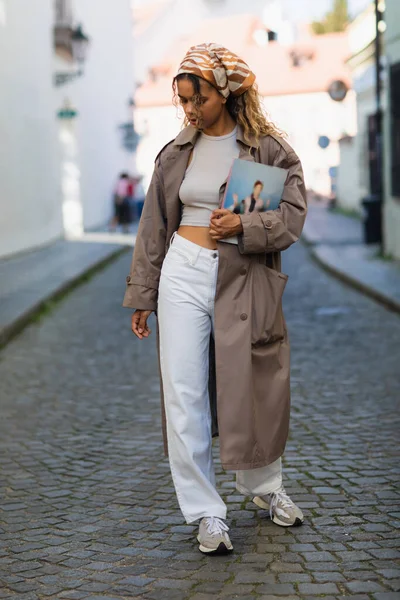 Full length of african american woman in headscarf and trench coat walking with magazine on street in prague — Fotografia de Stock