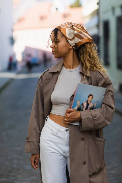 African american woman in headscarf and stylish outfit walking with magazine on street in prague - foto de stock