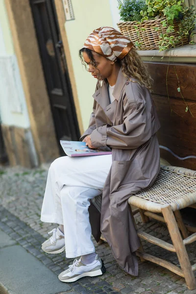 Stylish african american woman in headscarf and trench coat reading magazine on street in prague — Fotografia de Stock