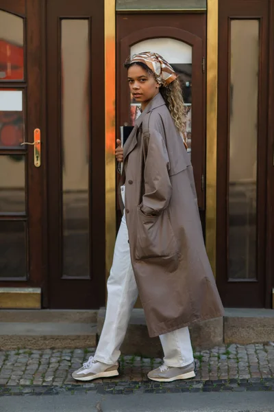 Full length of stylish african american woman in headscarf and trench coat walking with hand in pocket in prague — Photo de stock