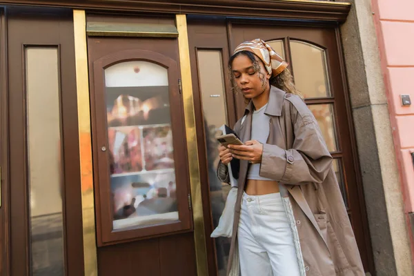 Stylish african american woman in headscarf and trench coat walking and using smartphone in prague — Fotografia de Stock