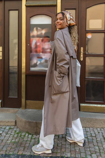 Full length of cheerful african american woman in headscarf and trendy trench coat walking with hand in pocket in prague - foto de stock