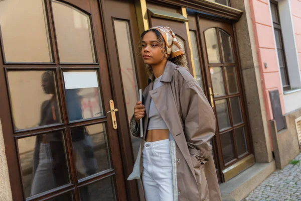 Stylish african american woman in headscarf and trench coat standing with hand in pocket near entrance door in prague — Foto stock