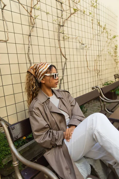 Stylish african american woman in headscarf and sunglasses sitting in trench coat on bench in prague — Stockfoto