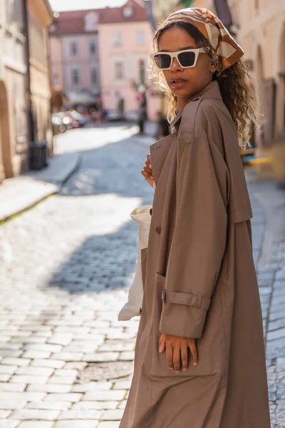 Stylish african american woman in headscarf and sunglasses walking in trench coat on street in prague — Stock Photo