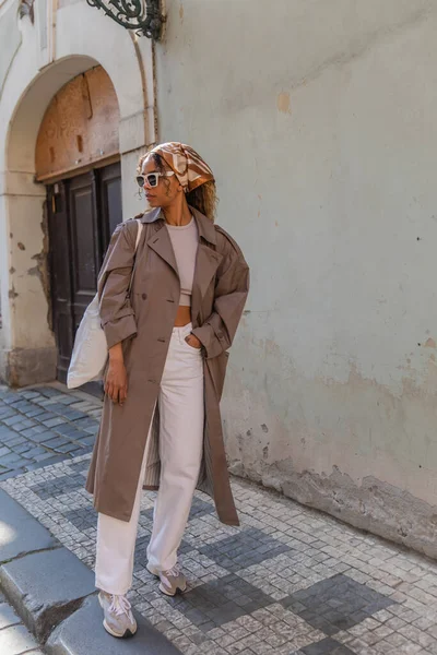 Полная длина стильная африканская американка в платке и солнцезащитных очках позирует с многоразовой сумкой на улице в Праге — стоковое фото