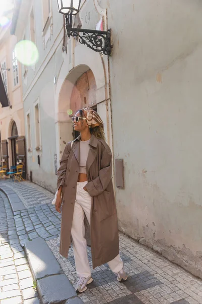 Full length of stylish african american woman in trendy accessories posing with hand in pocket on street in prague - foto de stock