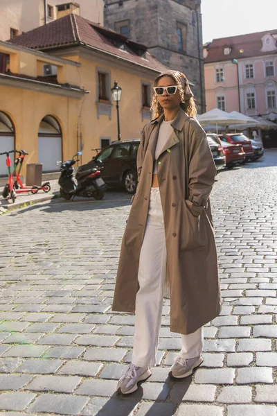 Full length of curly african american woman in stylish accessories posing with hand in pocket on street in prague - foto de stock