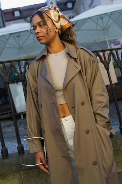 Young african american woman in stylish outfit and headscarf posing with hand in pocket on street in prague - foto de stock