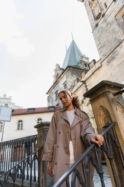 Low angle view of stylish african american woman in trench coat near old town hall tower in prague — Foto stock