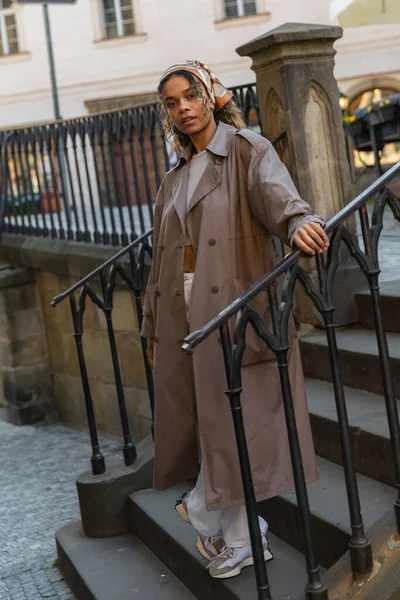 Full length of trendy african american woman in trench coat standing on stairs - foto de stock