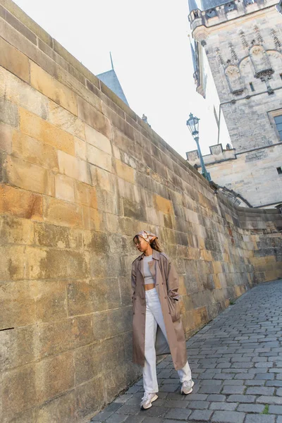 Full length of stylish african american woman in trench coat walking near ancient wall in prague — Stock Photo