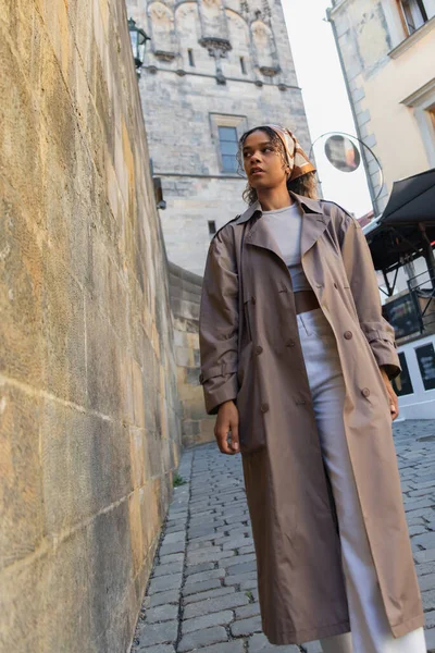 Low angle view of stylish african american woman in trench coat walking near ancient wall in prague — Stockfoto