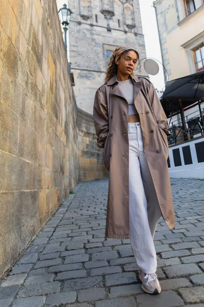Full length of stylish african american woman in trendy trench coat walking with hands in pockets near ancient wall in prague — Photo de stock