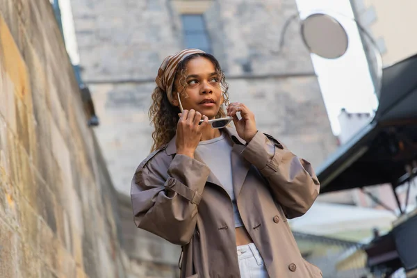 Low angle view of stylish african american woman in trendy trench coat wearing sunglasses in street in prague — Foto stock