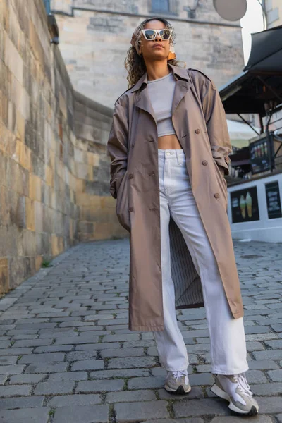 Full length of stylish african american woman in trendy trench coat and sunglasses posing on street in prague — Stock Photo