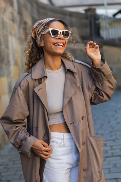Cheerful african american woman in trendy trench coat and sunglasses gesturing on street in prague — Photo de stock