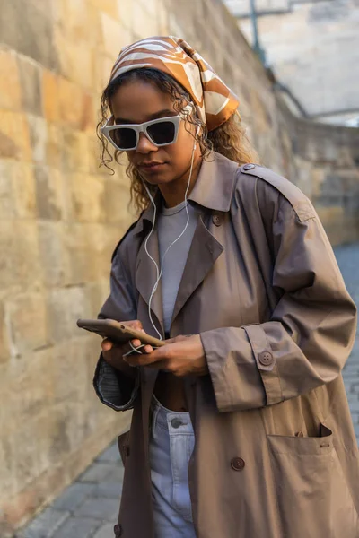 Stylish african american woman in trendy accessories listening music in wired earphones and using smartphone — Foto stock