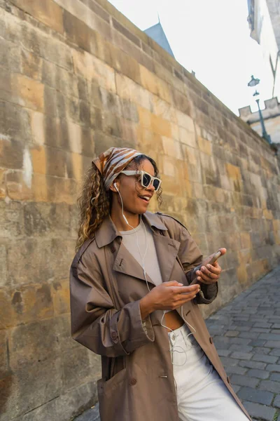 Happy african american woman in trendy accessories listening music in wired earphones and using smartphone — Photo de stock