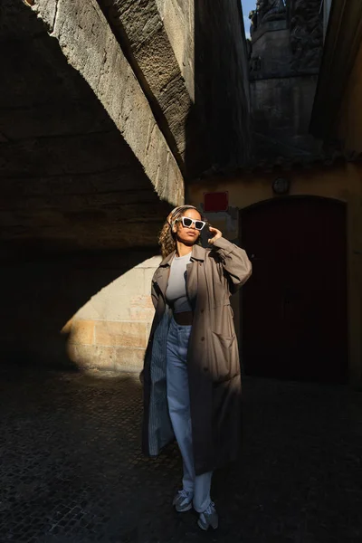 Stylish african american woman in trendy accessories and trench coat standing on spotlight from sunlight — Stock Photo