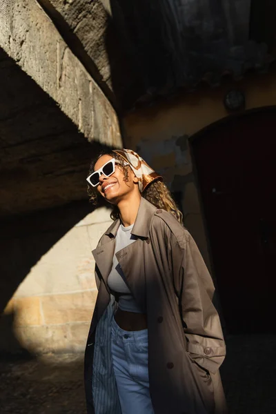 Happy african american woman in trendy accessories and trench coat standing on spotlight from sunlight — Fotografia de Stock