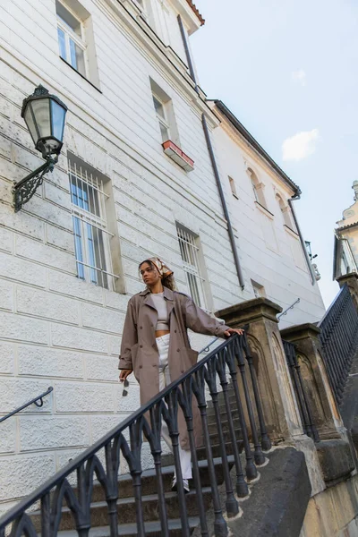 African american woman in trendy headscarf and trench coat touching handrail while walking on stairs in prague — Stockfoto