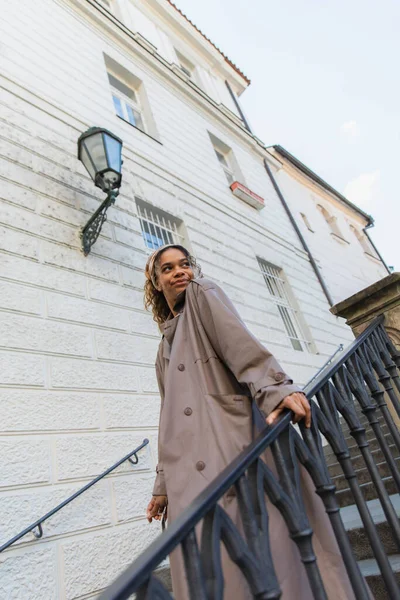 Pleased african american woman in trendy headscarf and trench coat touching handrail while walking on stairs in prague — Stock Photo
