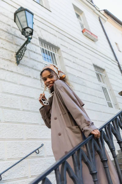 Low angle view of happy african american woman in trendy headscarf holding sunglasses and standing on stairs in prague — Foto stock