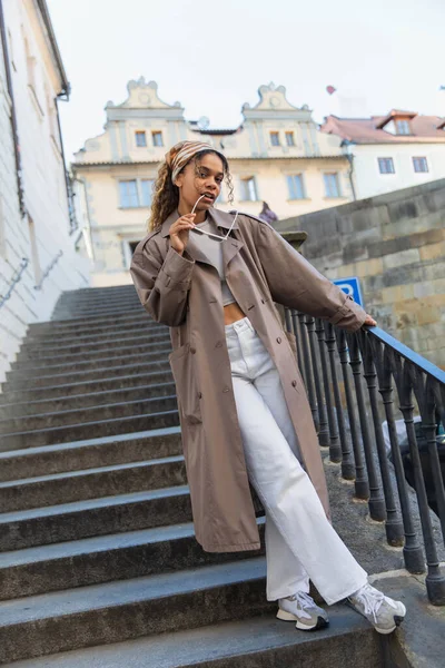 Young african american woman in trendy headscarf holding sunglasses and standing on stairs in prague — Foto stock