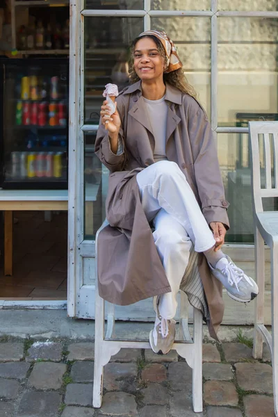 Весёлая африканская американка в платке и стильном плаще держит мороженое и сидит на стуле в Праге — стоковое фото