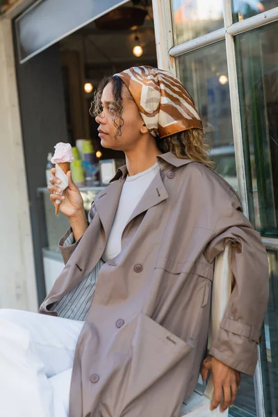 Young african american woman in headscarf and stylish trench coat holding ice cream cone and sitting on chair in prague - foto de stock