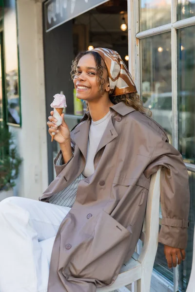 Joyful african american woman in headscarf and stylish trench coat holding ice cream cone and sitting on chair in prague — Fotografia de Stock